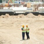 Two workers discussing plans on a sandy construction site, wearing safety gear.